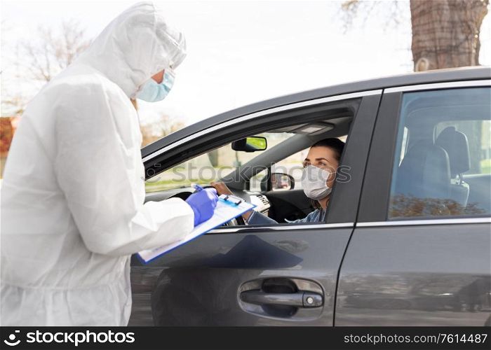 medicine, quarantine and pandemic concept - doctor or healthcare worker in protective gear, medical mask, gloves and goggles with clipboard and woman waiting for coronavirus test in her car. healthcare worker with clipboard and woman in car