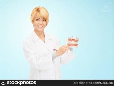 medicine, stomatology, people and dental hygiene concept - smiling female doctor with toothbrush and jaws model teaching how to brush teeth over blue background