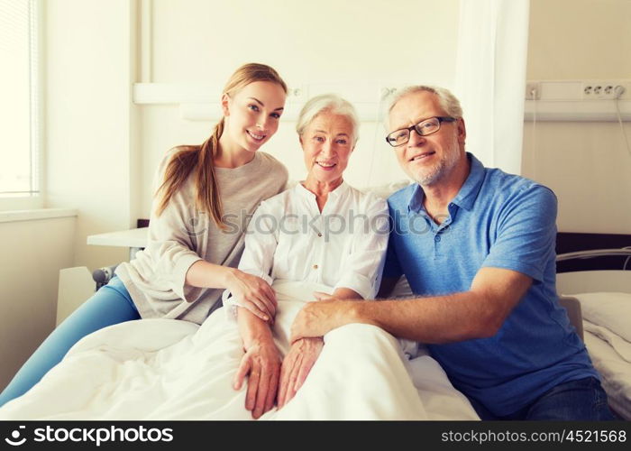 medicine, support, family health care and people concept - happy senior man and young woman visiting and cheering her grandmother lying in bed at hospital ward