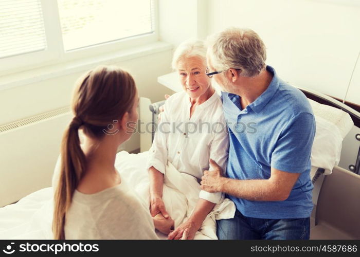 medicine, support, family health care and people concept - happy senior man and young woman visiting and cheering her grandmother lying in bed at hospital ward