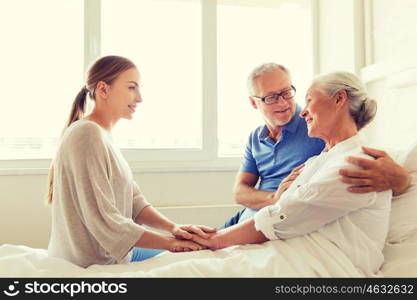 medicine, support, family health care and people concept - happy senior man and young woman visiting and cheering her grandmother lying in bed at hospital ward