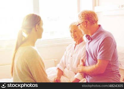 medicine, support, family health care and people concept - happy senior man and young woman visiting and cheering her grandmother lying in bed at hospital ward