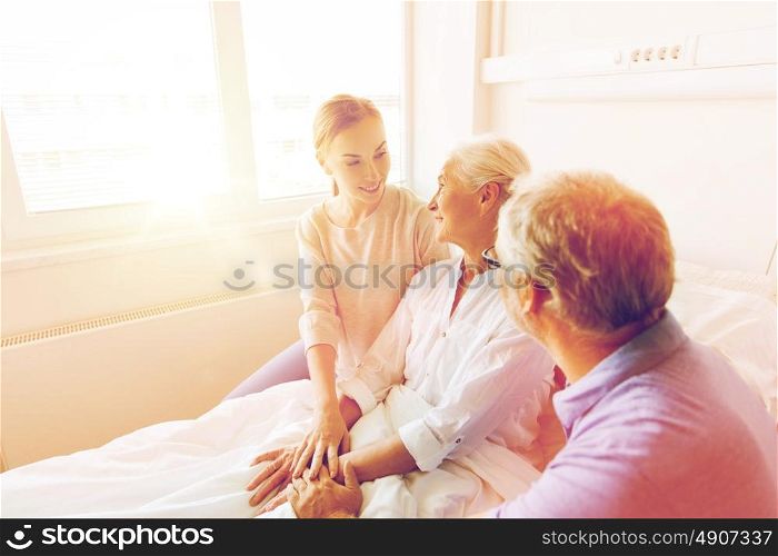 medicine, support, family health care and people concept - happy senior man and young woman visiting and cheering her grandmother lying in bed at hospital ward. happy family visiting senior woman at hospital