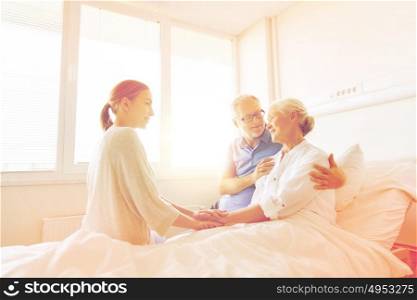 medicine, support, family health care and people concept - happy senior man and young woman visiting and cheering her grandmother lying in bed at hospital ward. happy family visiting senior woman at hospital