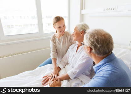 medicine, support, family health care and people concept - happy senior man and young woman visiting and cheering her grandmother lying in bed at hospital ward
