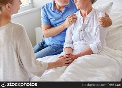 medicine, support, family health care and people concept - senior man and young woman visiting and cheering her grandmother lying in bed at hospital ward. family visiting senior woman at hospital