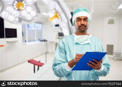 medicine, surgery and healthcare concept - smiling indian male doctor or surgeon in protective wear with clipboard over operating room at hospital background. indian male surgeon with clipboard at hospital