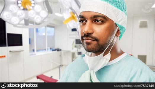 medicine, surgery and people concept - close up of indian male doctor or surgeon with protective mask over operating room in hospital background. indian male doctor or surgeon at hospital