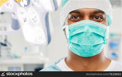 medicine, surgery and people concept - close up of indian male doctor or surgeon in mask and protective wear over operating room at hospital background. indian male surgeon in mask over operating room