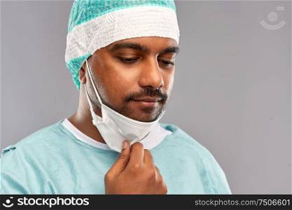 medicine, surgery and people concept - close up of sad indian male doctor or surgeon with protective mask over grey background. face of sad doctor or surgeon with protective mask