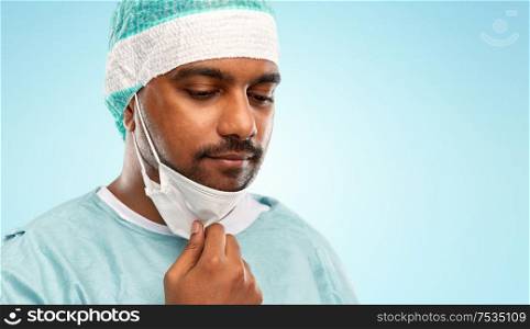 medicine, surgery and people concept - close up of sad indian male doctor or surgeon with protective mask over blue background. face of sad doctor or surgeon with protective mask