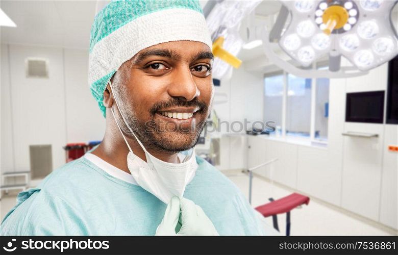 medicine, surgery and people concept - close up of smiling indian male doctor or surgeon with protective mask over operating room in hospital background. smiling indian male doctor or surgeon at hospital