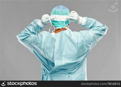 medicine, surgery and people concept - indian male doctor or surgeon in protective wear putting face mask on over grey background. indian male doctor or surgeon in protective wear
