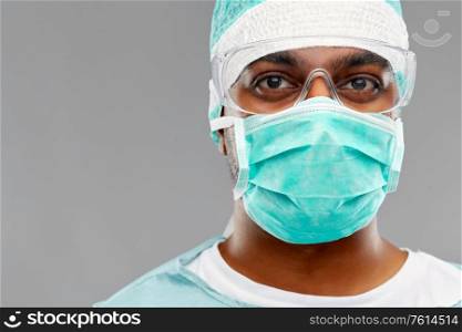medicine, surgery and people concept - indian male doctor or surgeon in mask, goggles and protective wear over grey background. indian male doctor or surgeon in protective wear
