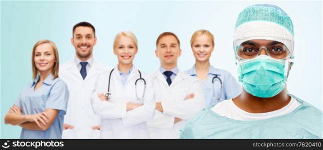 medicine, surgery and people concept - indian male doctor or surgeon in mask, goggles and protective wear over smiling healthcare workers team on blue background. indian doctor or surgeon and healthcare workers