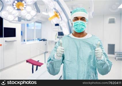medicine, surgery and people concept - indian male doctor or surgeon in mask and protective wear showing thumbs up over operating room at hospital background. indian male doctor or surgeon over operating room