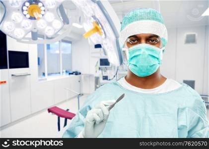 medicine, surgery and people concept - indian male doctor or surgeon in mask and protective wear with scalpel knife over operating room at hospital background. indian surgeon with scalpel over operating room
