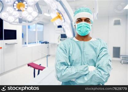 medicine, surgery and people concept - indian male doctor or surgeon in mask and protective wear with crossed arms over operating room at hospital background. indian male doctor or surgeon over operating room