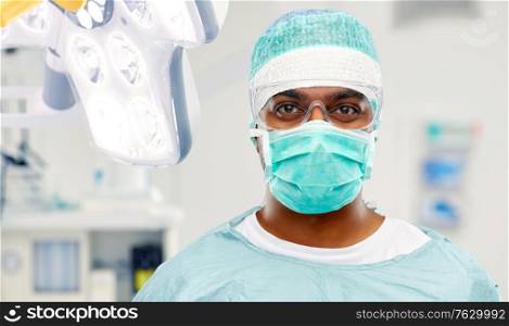 medicine, surgery and people concept - indian male doctor or surgeon in mask, goggles and protective wear over operating room at hospital background. indian male surgeon in mask over operating room