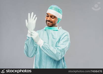 medicine, surgery and people concept - smiling indian male doctor or surgeon in protective wear putting glove on over grey background. indian male doctor or surgeon putting glove on