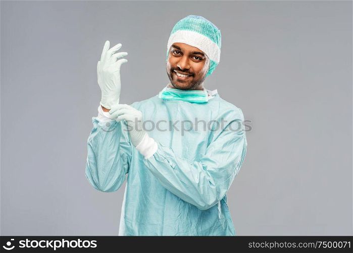 medicine, surgery and people concept - smiling indian male doctor or surgeon in protective wear putting glove on over grey background. indian male doctor or surgeon putting glove on
