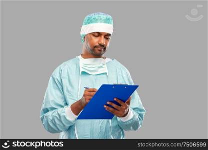 medicine, surgery and people concept - smiling indian male doctor or surgeon in protective wear with clipboard over grey background. indian male doctor or surgeon with clipboard