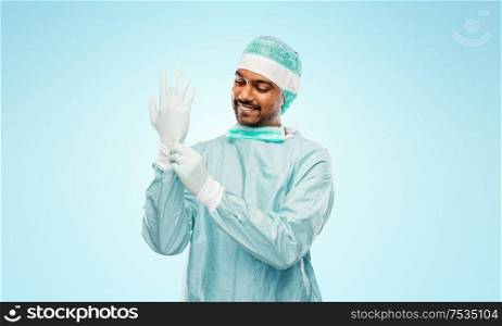 medicine, surgery and people concept - smiling indian male doctor or surgeon in protective wear putting glove on over grey background. indian male doctor or surgeon putting glove on