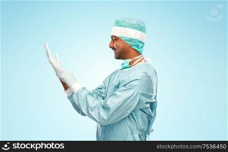 medicine, surgery and people concept - smiling indian male doctor or surgeon in protective wear putting glove on over blue background. indian male doctor or surgeon putting glove on