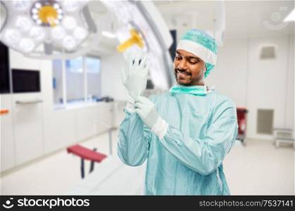 medicine, surgery and people concept - smiling indian male doctor or surgeon in protective wear putting glove on over operating room in hospital background. indian male doctor or surgeon at hospital
