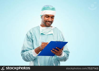 medicine, surgery and people concept - smiling indian male doctor or surgeon in protective wear with clipboard over blue background. indian male doctor or surgeon with clipboard