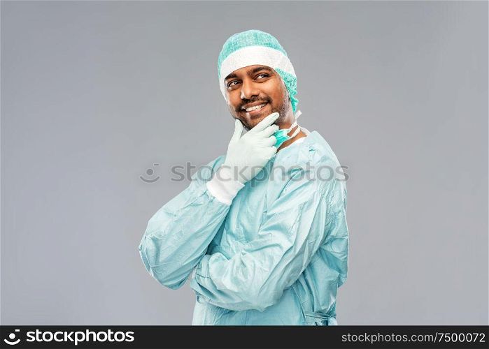 medicine, surgery and people concept - smiling thiniking indian male doctor or surgeon in protective wear over grey background. indian male doctor or surgeon in protective wear