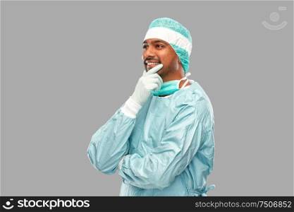 medicine, surgery and people concept - smiling thinking indian male doctor or surgeon in protective wear over grey background. indian male doctor or surgeon in protective wear