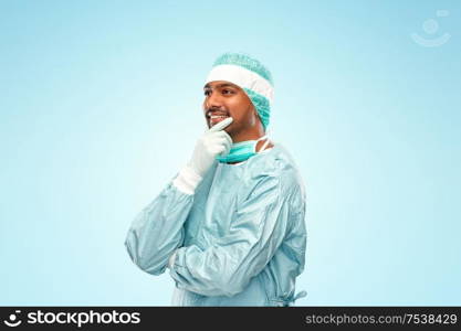 medicine, surgery and people concept - smiling thinking indian male doctor or surgeon in protective wear over blue background. indian male doctor or surgeon in protective wear