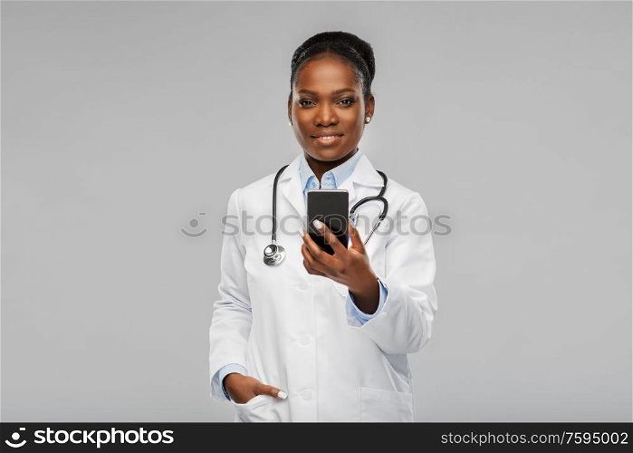 medicine, technology and healthcare concept - happy smiling african american female doctor or in white coat with smartphone and stethoscope over background. african american female doctor with smartphone