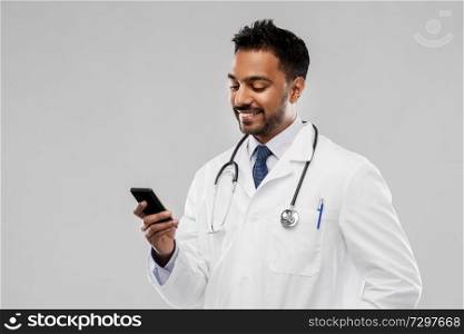 medicine, technology and healthcare concept - smiling indian male doctor in white coat with smartphone and stethoscope over grey background. smiling indian male doctor with smartphone