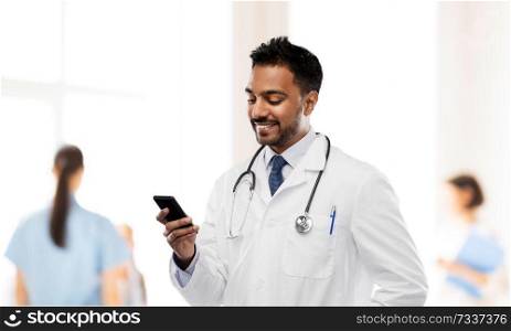 medicine, technology and healthcare concept - smiling indian male doctor in white coat with smartphone and stethoscope over hospital background. smiling indian male doctor with smartphone