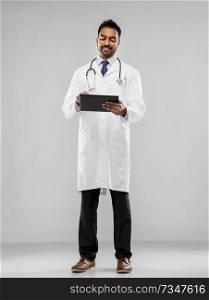 medicine, technology and healthcare concept - smiling indian male doctor in white coat with tablet computer and stethoscope over grey background. smiling indian male doctor with tablet computer