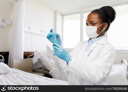 medicine, vaccination and healthcare concept - african american female doctor in mask with syringe over hospital ward background. african american doctor with syringe at hospital