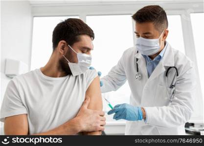 medicine, vaccination and healthcare concept - doctor wearing face protective medical mask for protection from virus disease with syringe doing injection of vaccine to male patient. male doctor in mask giving vaccine to patient