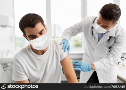 medicine, vaccination and healthcare concept - doctor with syringe doing injection of vaccine to male patient wearing face protective medical masks for protection from virus disease. patient and doctor in masks doing vaccination