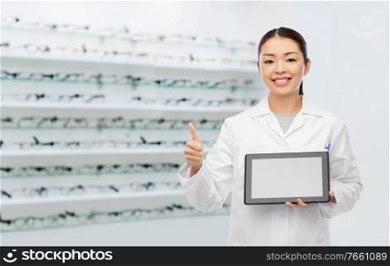 medicine, vision and healthcare concept - happy smiling asian female eye doctor or ophthalmologist with tablet pc computer showing thumbs up over glasses at optical store background. asian female doctor with tablet pc shows thumbs up