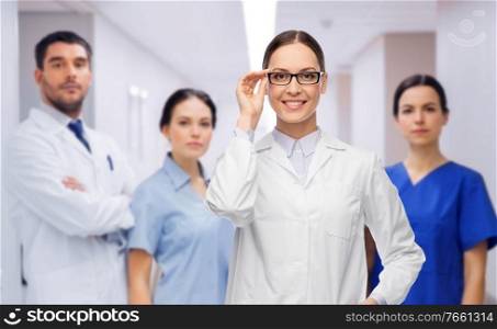 medicine, vision and healthcare concept - happy smiling female doctor in glasses and white coat over colleagues at hospital on background. smiling female doctor with colleagues at hospital