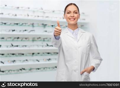 medicine, vision and healthcare concept - happy smiling female eye doctor or ophthalmologist in white coat showing thumbs up over glasses at optical store background. female doctor showing thumbs up at optical store