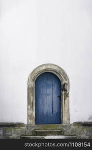 Medieval arched door. Aged wooden door in blue color with stone stairs and white wall. Just one door and an empty white house wall. Old blue entrance.