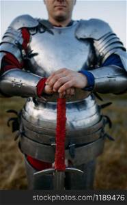 Medieval knight in metal armor holds sword, great battle. Armored ancient warrior in armour posing in the field