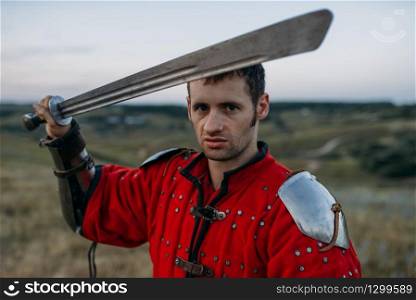 Medieval knight in metal helmet holds sword, great battle. Armored ancient warrior in armour posing in the field. Medieval knight in metal helmet holds sword