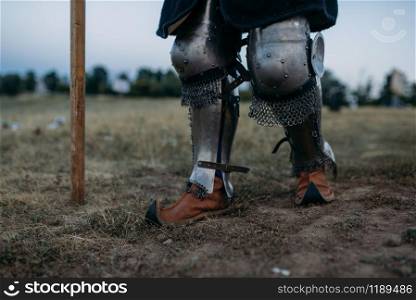 Medieval knight legs in metal armor, back view, great tournament. Armored ancient warrior in armour posing in the field. Medieval knight legs in metal armor, back view