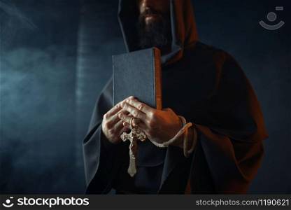 Medieval monk holds book and wooden cross in hands, black background, secret ritual. Mysterious friar in dark cape. Mystery and spirituality. Medieval monk holds book and wooden cross in hands