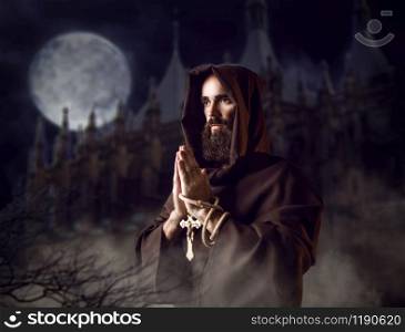 Medieval monk in black robe with hood praying against castle and full moon in the night, secret ritual. Mysterious friar in dark cape. Mystery and spirituality