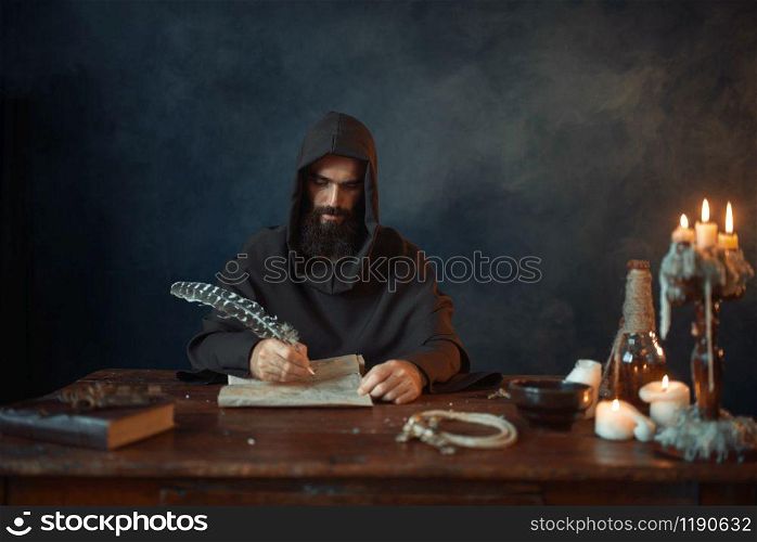 Medieval monk in robe and hood writes with a goose feather, black background, secret ritual. Mysterious friar in dark cape. Mystery and spirituality. Medieval monk in robe writes with a goose feather